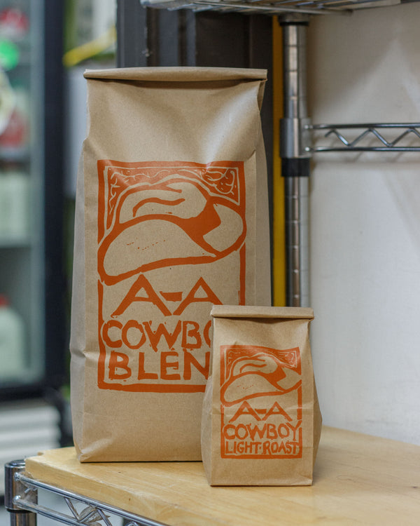 Two brown paper bags with A-A Cowboy Blend logo on a wooden surface.