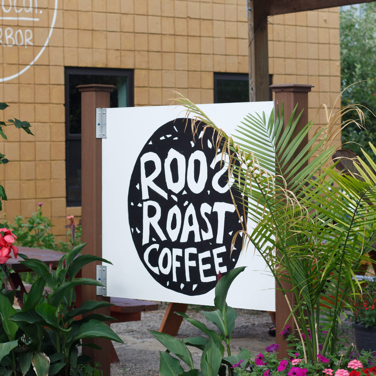 A white outdoor sign for Roo's Roast Coffee with a large black circular logo, displayed on a stand in front of a tan building with green plants and flowers.