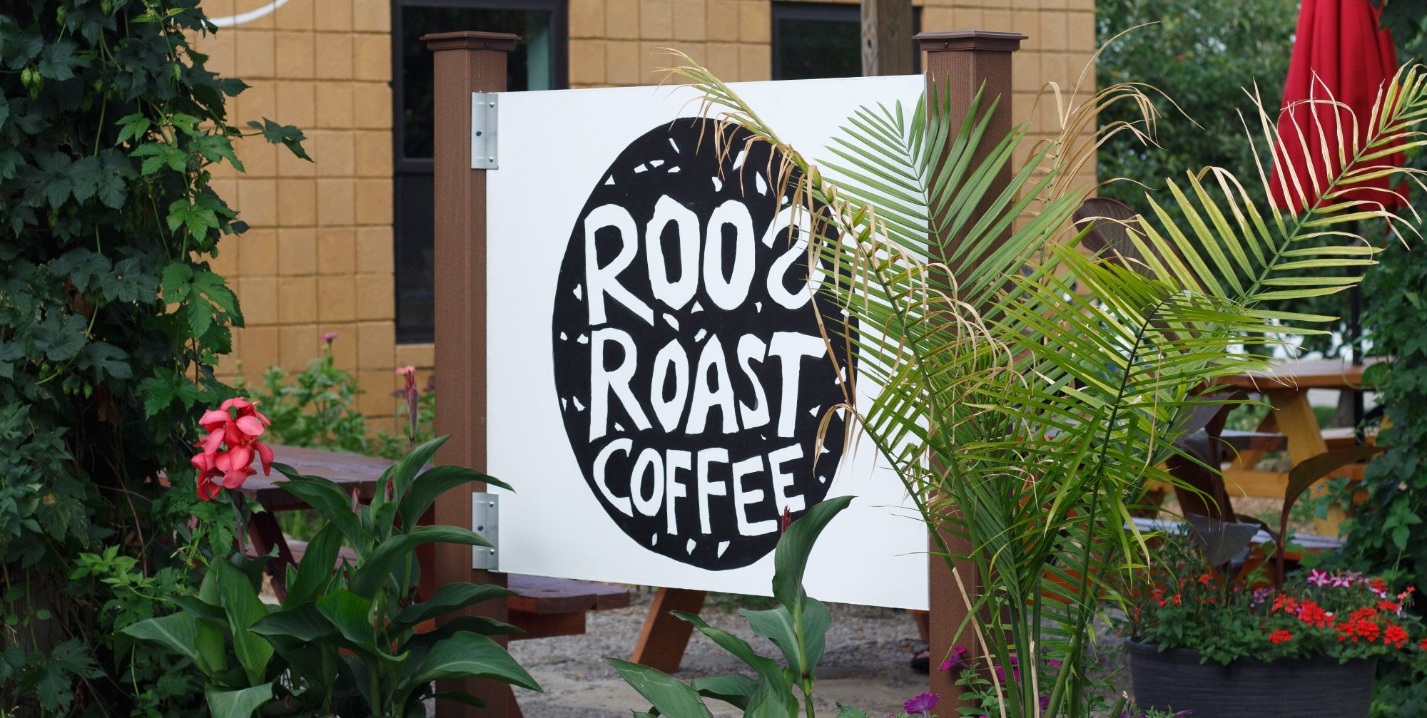 A white outdoor sign for Roo's Roast Coffee with a large black circular logo, displayed on a stand in front of a tan building with green plants and flowers.