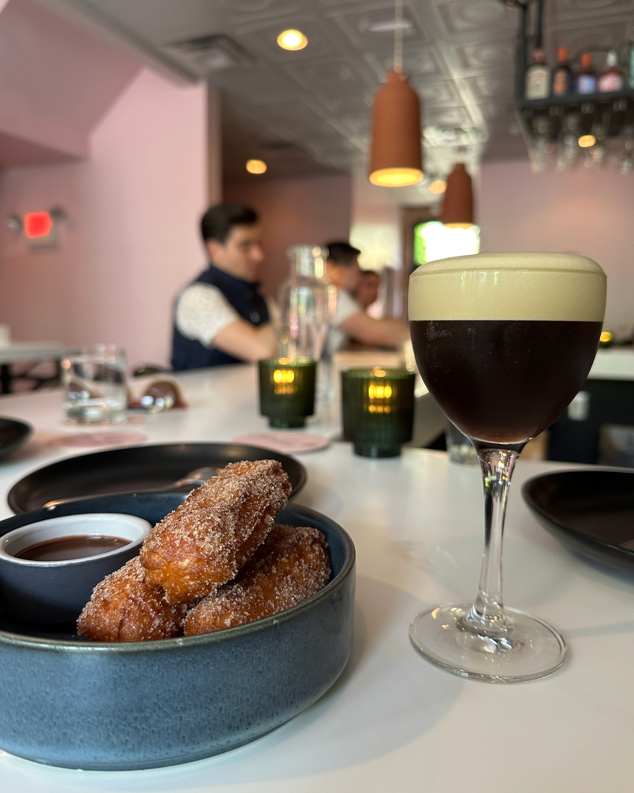 Café setting with a glass of espresso and a bowl of fried pastries on a table.