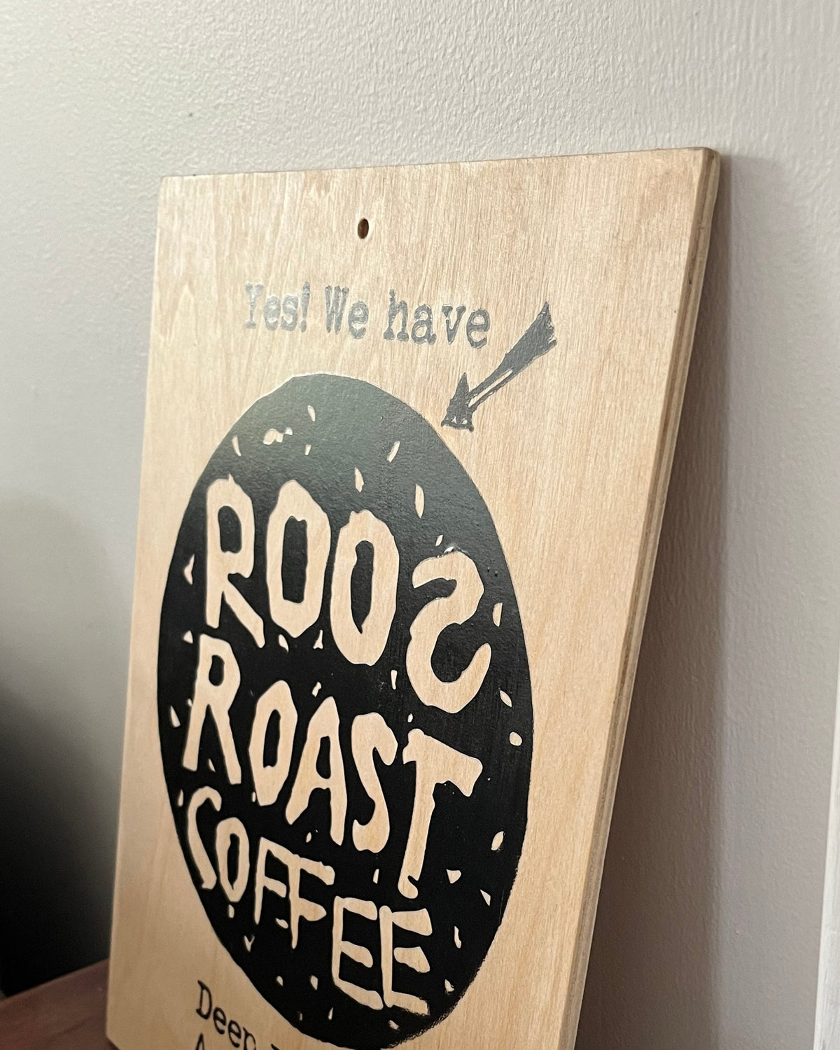 A rectangular wooden coffee sign leaning against a white wall on a dark wooden table. The sign has a natural light wood background with a large black oval in the center containing the words 'ROO2 ROAST COFFEE' in cream lettering, and smaller text above and below.