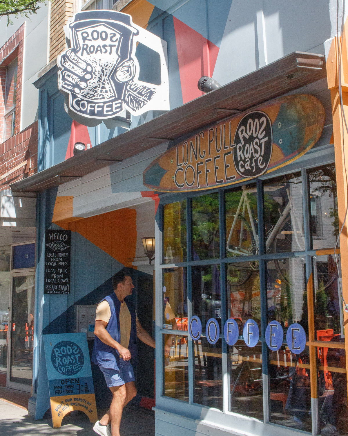 A storefront scene for Root Roast Coffee on a sunny street. The shop has a colorful painted facade with blue, orange, and white sections, large glass windows, and multiple visible signs including a large circular logo above the entrance and a smaller sign on the sidewalk.