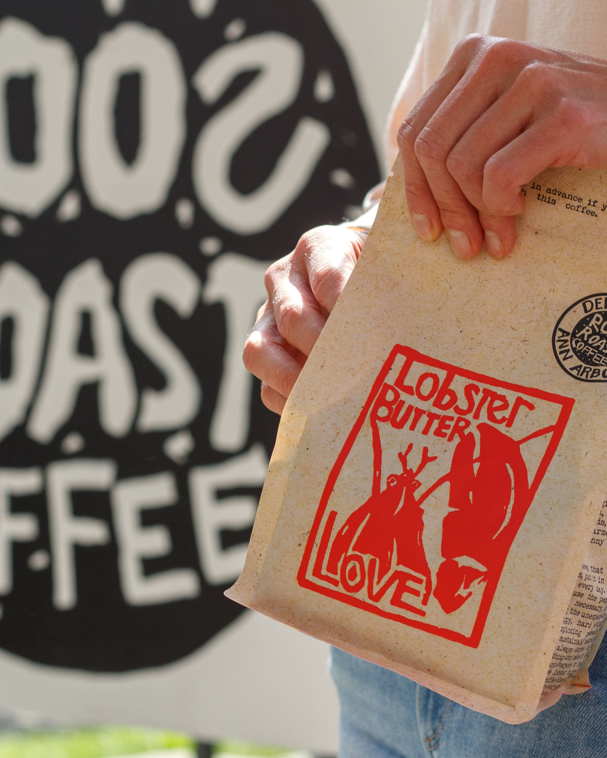 Person holding a coffee package with 'Lobster Butter Love' design in front of a 'RoosRoast Coffee' sign.