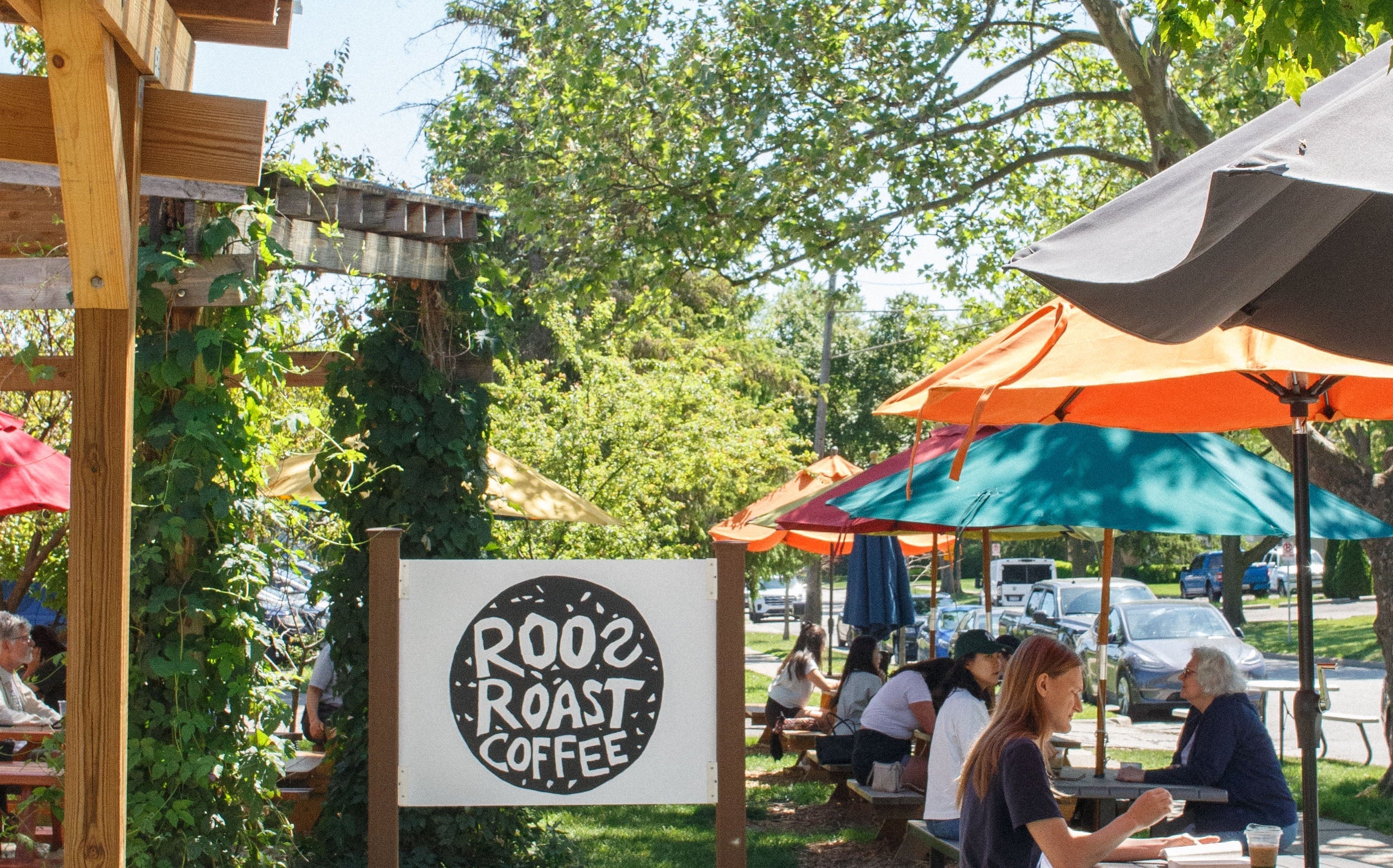 Outdoor seating area with people at tables under colorful umbrellas, featuring a 'Roo's Roast Coffee' sign.