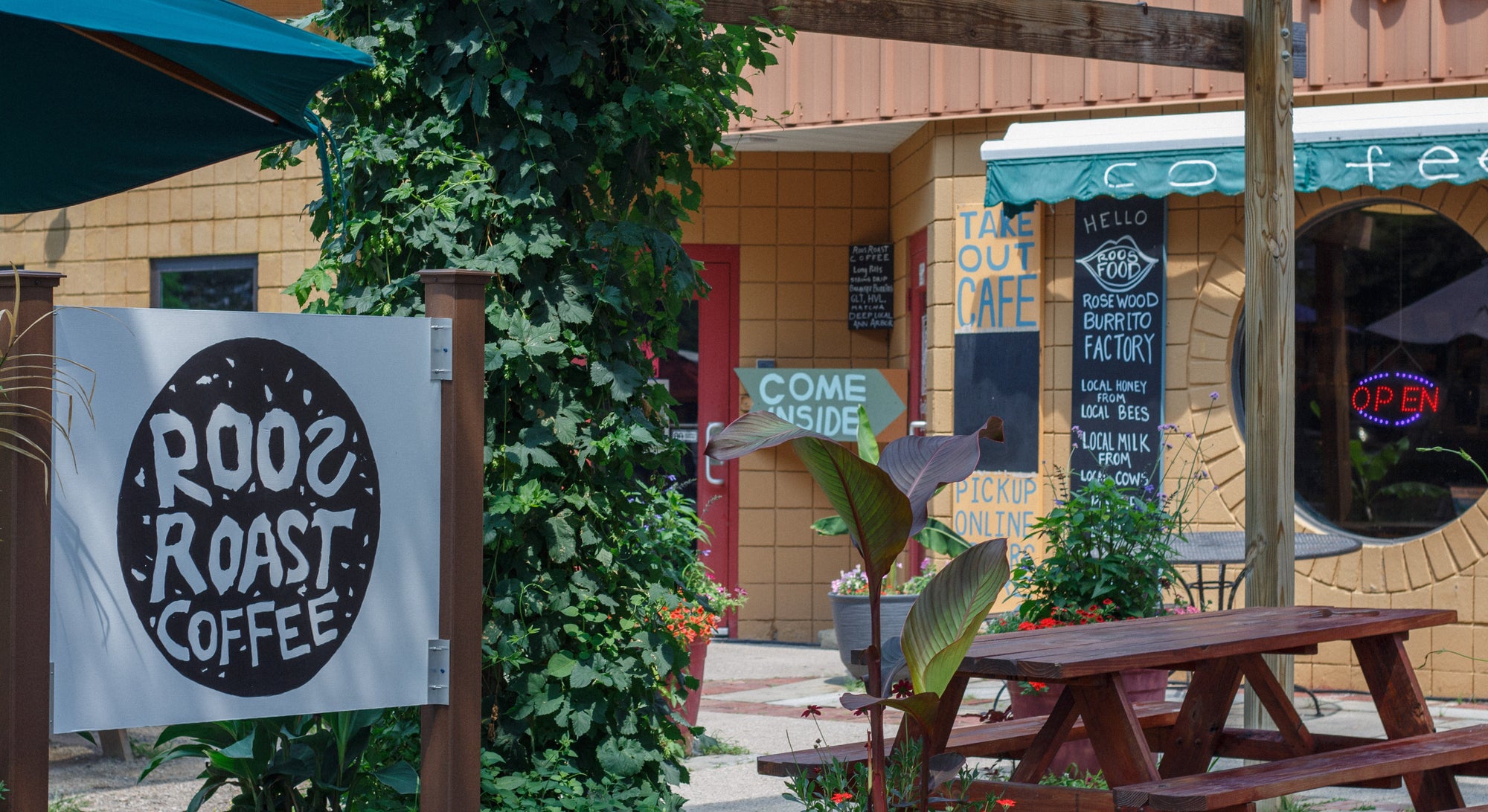 Outdoor storefront scene for Roost Roast Coffee, with a white sign reading 'ROOST ROAST COFFEE' on a wooden post, greenery, picnic tables, and a tan building facade in the background.