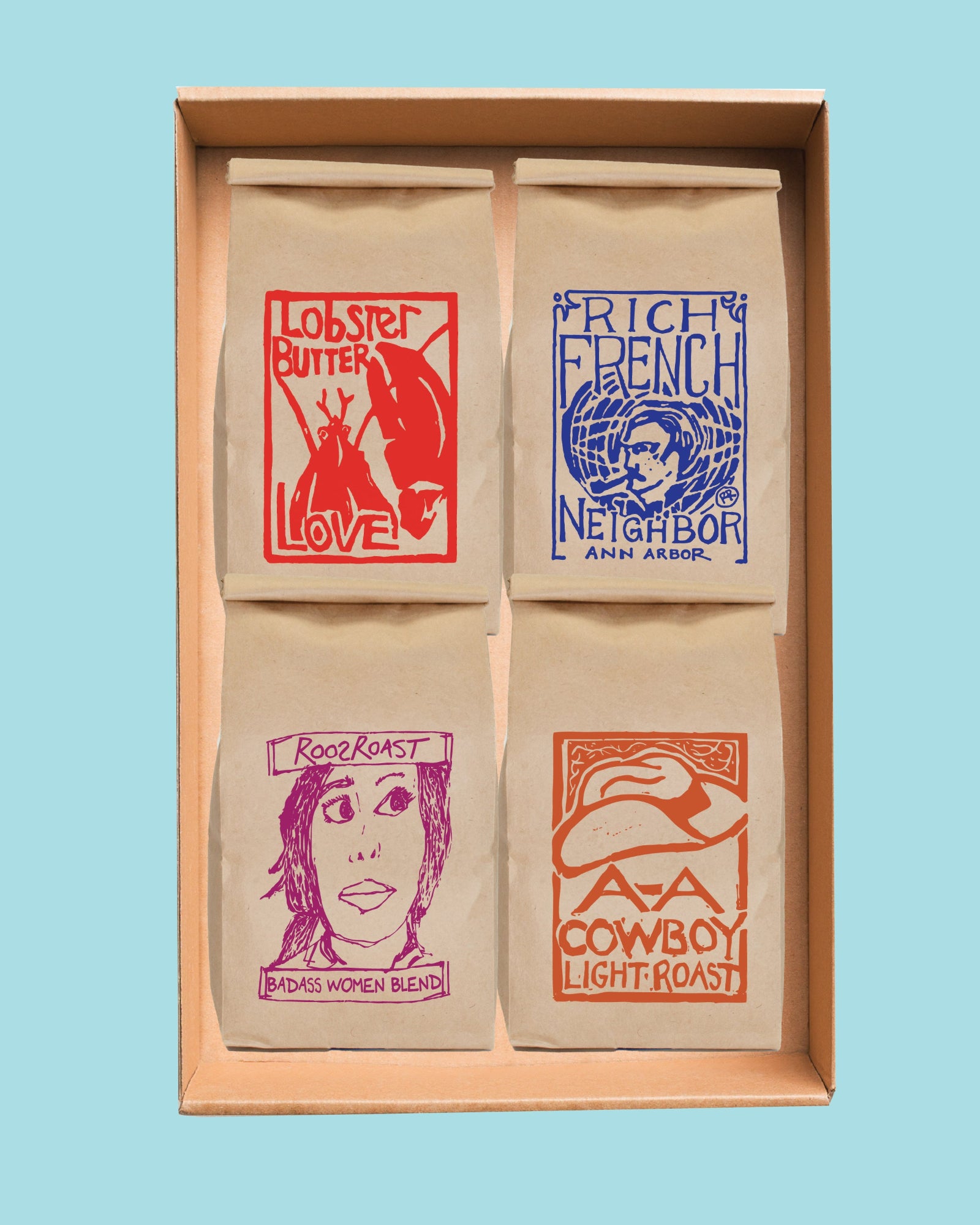 A boxed set of four paper coffee bags arranged in a 2x2 grid inside a light wood tray, each bag printed with a different graphic and text including 'Lobster Butter Love,' 'Rich French Neighbor,' 'Rooster,' and 'AA Cowboy Light Roast,' on a light blue background.