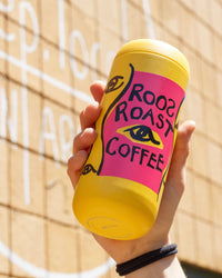 Yellow and pink coffee cup with 'Roo's Roast Coffee' branding held against a textured wall.